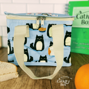 Lunch bag on a wooden surface with a cream handle from the bag draped in front of it. The bag is light blue with black and white cats on it.