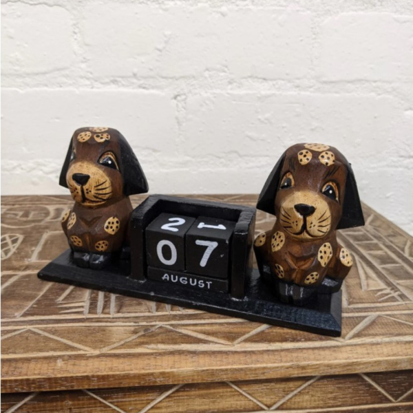 Hand-carved wooden dog perpetual calendar with two brown dogs and black date blocks showing “07 August,” displayed on a patterned wooden table with a white brick wall background.