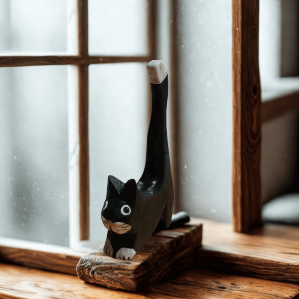 Black hand-carved wooden cat ornament with a tall tail and white-tipped detail, shown on a wooden windowsill with soft natural light. A charming decorative piece for cat lovers.