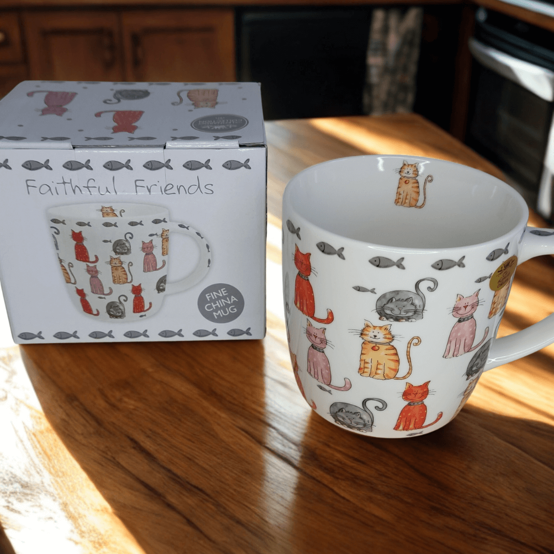 Faithful Friends cat-themed fine china mug positioned beside its matching gift box on a sunlit wooden table. The design includes orange, grey, and pink cats surrounded by small grey fish.
