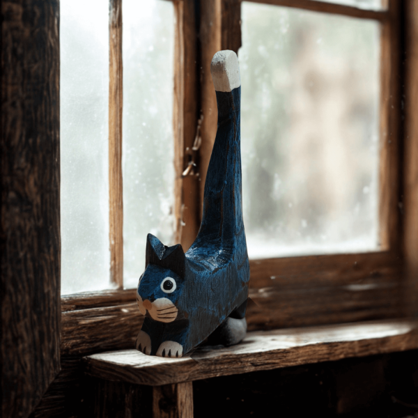 Blue wooden cat ornament with white facial details and a raised tail, placed on a rustic wooden windowsill. A hand-carved cat figure with a playful, folk-art style.