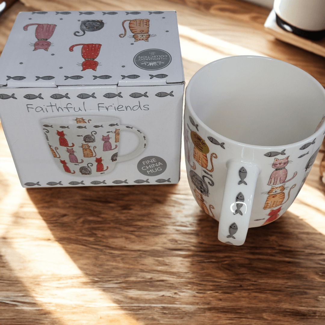Faithful Friends fine china cat mug and matching gift box displayed on a wooden table in natural sunlight. The mug and box both feature colourful cat illustrations with grey fish accents.