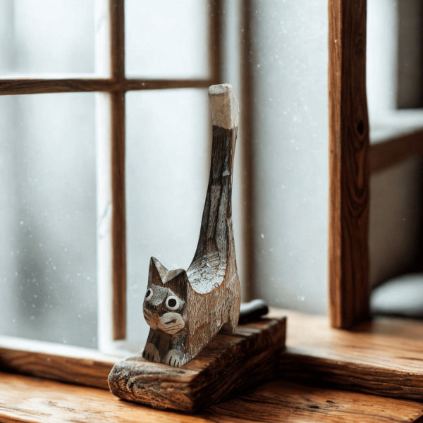 Hand-carved wooden cat ornament in natural wood tones displayed on a rustic windowsill. The cat features a raised tail and painted facial details, adding a cosy handmade charm to the scene.