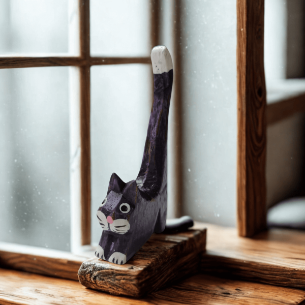 Purple hand-carved wooden cat ornament with painted details and a tall white-tipped tail, displayed on a windowsill in soft natural light. A whimsical handmade decorative figure.