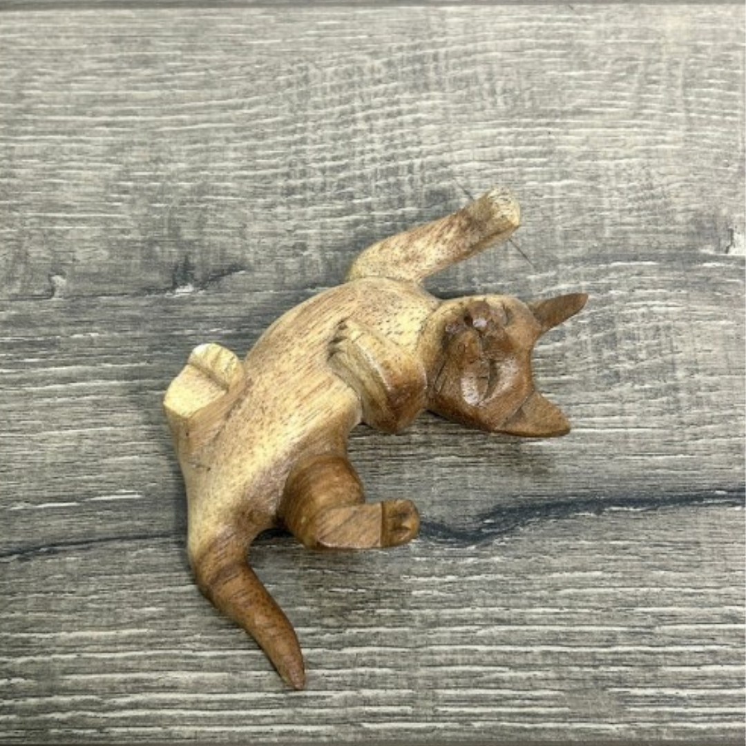 Hand-carved wooden cat ornament in a relaxed, playful position, displayed on a wooden background. Each piece is unique and handmade in Bali.