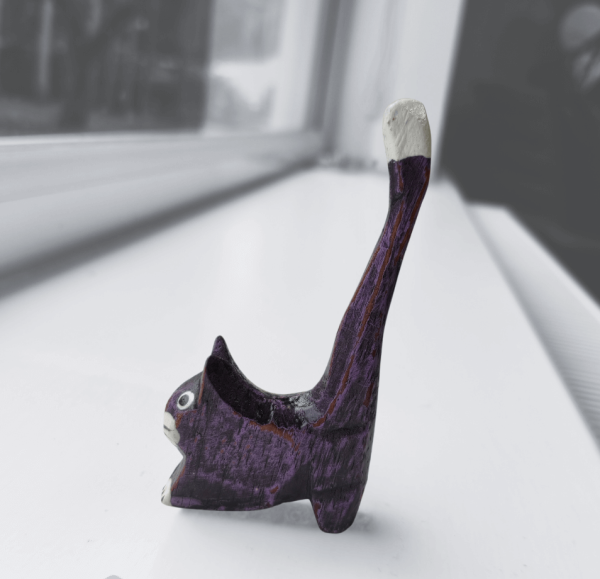 Purple hand-painted wooden cat ornament with a white tail tip and expressive eyes, photographed on a bright white windowsill. A quirky handmade décor piece for any home.