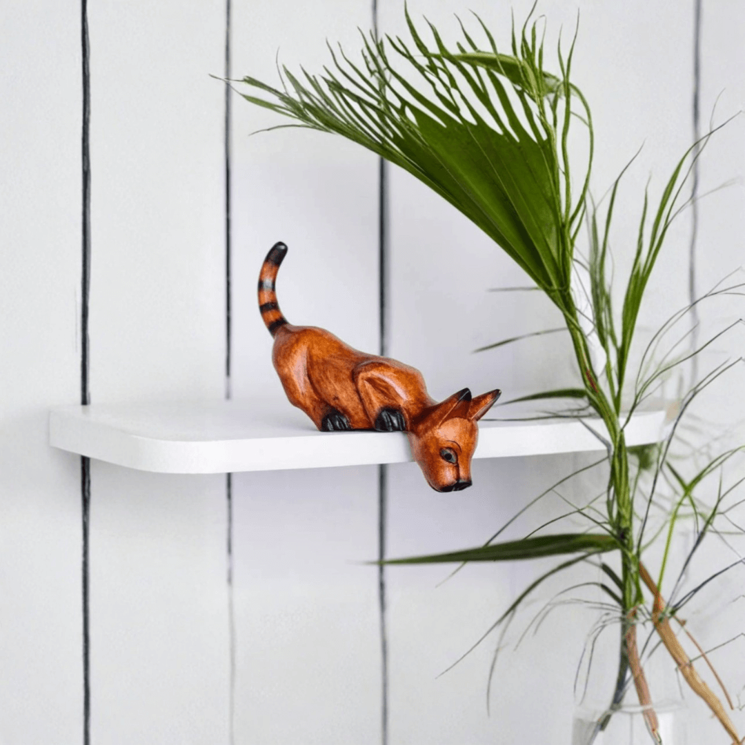 Wooden Shelf Sitter Cat ornament leaning over the edge of a white shelf with tall green plants beside it.