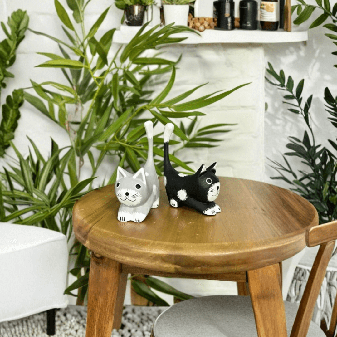 Set of two hand-carved wooden stretching cat ornaments in black and grey, displayed together on a wooden stool with indoor plants behind.