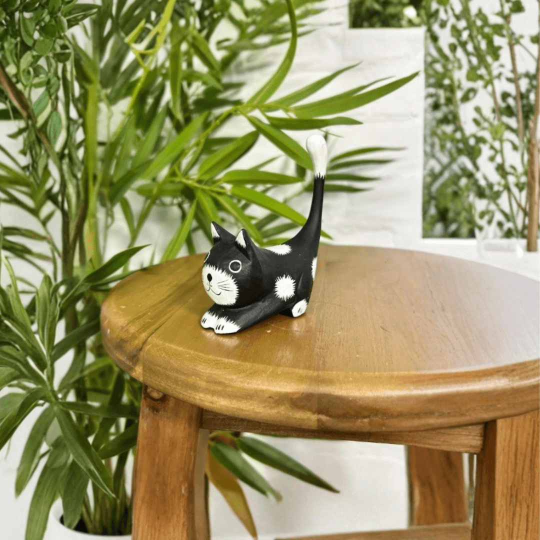 Hand-carved black wooden stretching cat ornament on a wooden stool surrounded by leafy green houseplants.