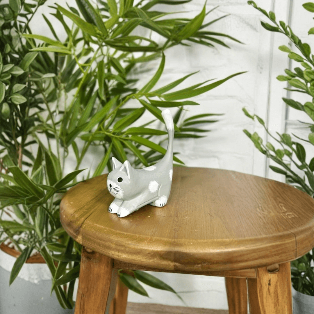 Hand carved grey wooden stretching cat ornament displayed on a wooden stool with green indoor plants in the background.