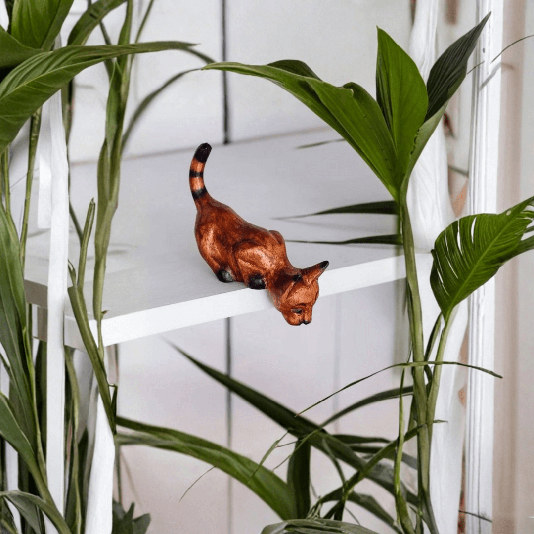 Hand-carved wooden Shelf Sitter Cat in a pounce position displayed on a white shelf with green houseplants around.