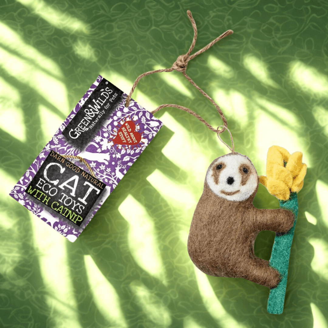 Handmade wool-felt sloth cat toy holding a yellow felt flower, attached to a jute string with a Green & Wilds tag, photographed on a green leafy background with soft sunlight shadows..
